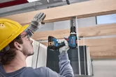 Một người mặc đồ bảo hộ đang khoan vít vào một thanh gỗ.