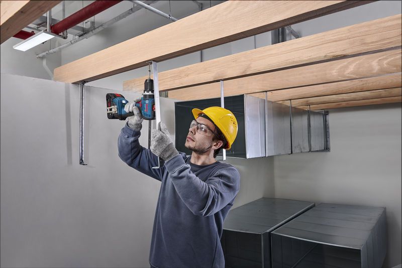 Một người mặc đồ bảo hộ đang khoan vào thanh kim loại bên dưới những thanh gỗ.