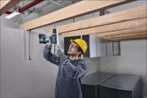 Một người mặc đồ bảo hộ đang khoan vào thanh kim loại bên dưới những thanh gỗ.