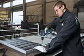 Một người mặc đồ bảo hộ đang khoan lưới kim loại trên bàn làm việc trong một xưởng.