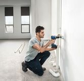 Một người đang khoan vào bức tường bên cạnh đường ống nước trong một căn phòng đang hoàn thiện một phần.