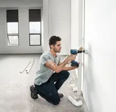 Một người đang khoan vào bức tường bên cạnh đường ống nước trong một căn phòng đang hoàn thiện một phần.