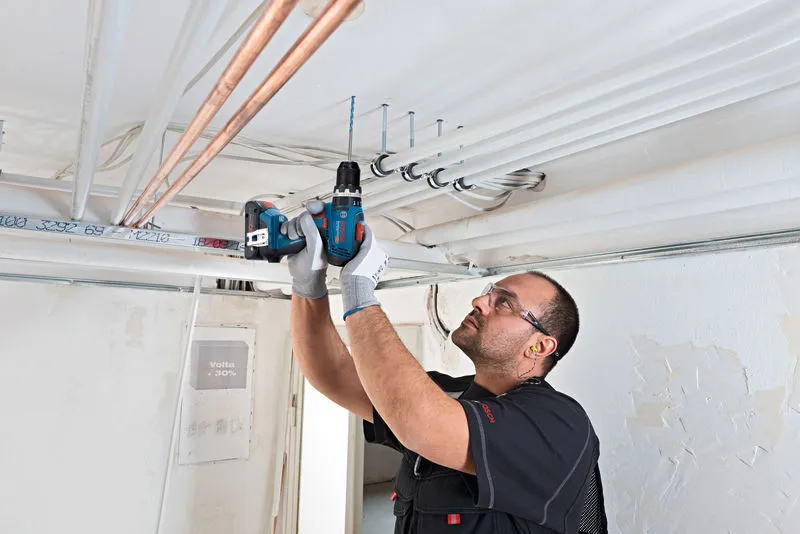 GSB 18V-65 Một người mặc thiết bị an toàn đang khoan vào trần nhà để lắp đặt ống dẫn điện.