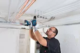 Một người mặc thiết bị an toàn đang khoan vào trần nhà để lắp đặt ống dẫn điện.
