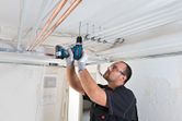 Một người mặc thiết bị an toàn đang khoan vào trần nhà để lắp đặt ống dẫn điện.