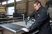 Một người mặc đồ bảo hộ đang khoan lưới kim loại trên bàn làm việc trong một nhà máy.