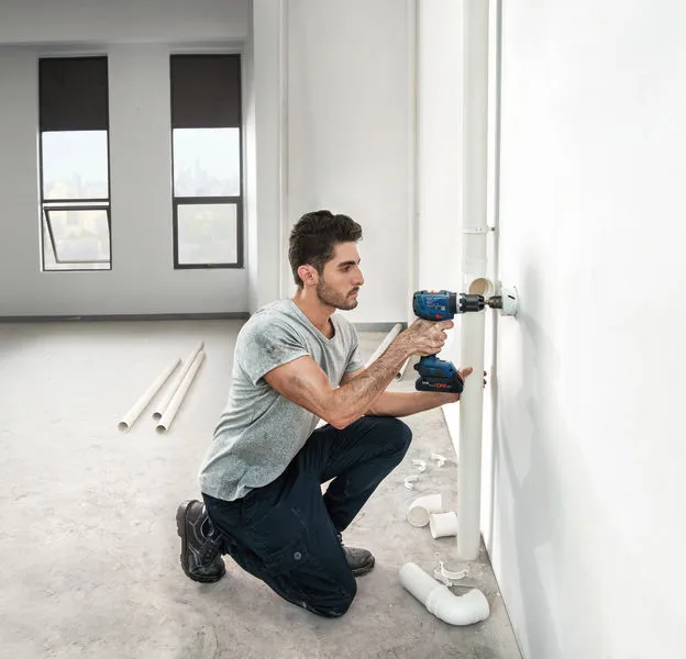 Một người đang khoan vào tường để lắp đường ống trong một căn phòng sáng sủa, chưa hoàn thiện.