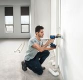 Một người đang khoan vào tường để lắp đường ống trong một căn phòng sáng sủa, chưa hoàn thiện.