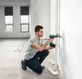 Một người đang khoan vào tường để lắp đường ống trong một căn phòng sáng sủa, chưa hoàn thiện.