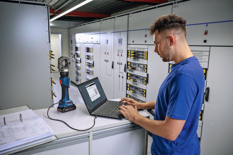 Người này đang vận hành máy tính xách tay được kết nối với dụng cụ bấm thủy lực không dây trong phòng điều khiển điện.