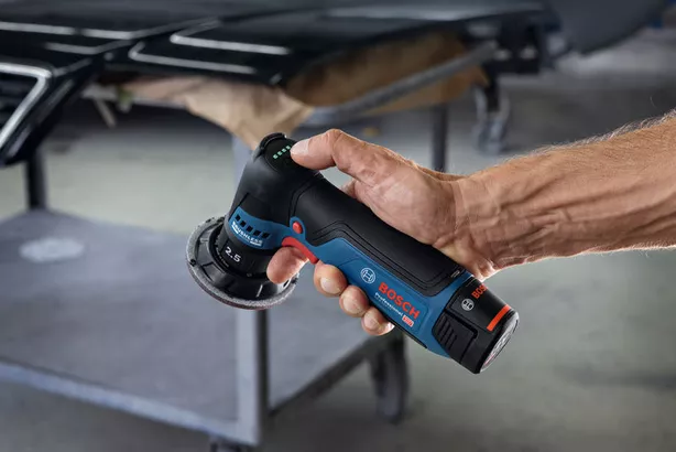 GEX 12V-77 Một người đang cầm máy chà nhám quỹ đạo ngẫu nhiên không dây bên cạnh bàn làm việc bằng kim loại.
