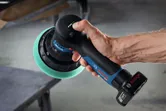 Một người đang cầm máy đánh bóng không dây có miếng đệm xốp màu xanh lá cây gần bàn làm việc.