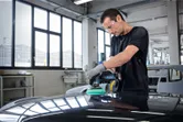 Một người mặc đồ bảo hộ đang đánh bóng mui xe ô tô bằng máy đánh bóng không dây.