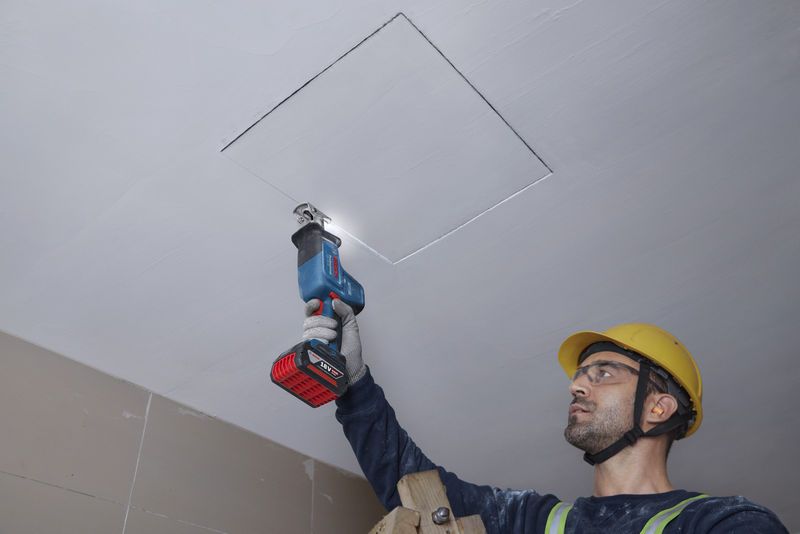 Một người mặc đồ bảo hộ đang cắt tấm trần nhà bằng máy cưa lọng.