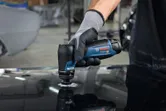 Một người mặc đồ bảo hộ đang chà nhám bề mặt kim loại bằng máy chà nhám quỹ đạo nhỏ gọn.