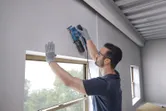 Một người mặc thiết bị an toàn đang làm phẳng bức tường bằng máy chà nhám quỹ đạo gần cửa sổ.