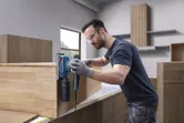 Một người mặc đồ bảo hộ đang chà nhám tủ gỗ bằng máy chà nhám quỹ đạo.