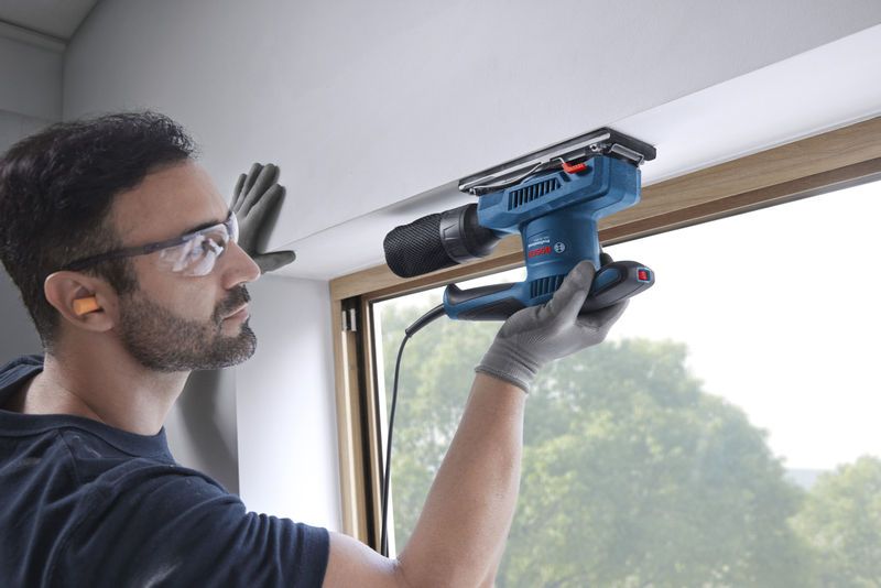 GSS 20-18 A Một người mặc đồ bảo hộ đang chà nhám khung cửa sổ bằng máy chà nhám quỹ đạo.