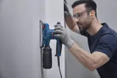 Một người mặc đồ bảo hộ đang sử dụng máy chà nhám quỹ đạo trên bức tường màu trắng.