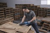 Một người mặc đồ bảo hộ đang cắt một tấm pallet gỗ bằng máy cưa lọng.