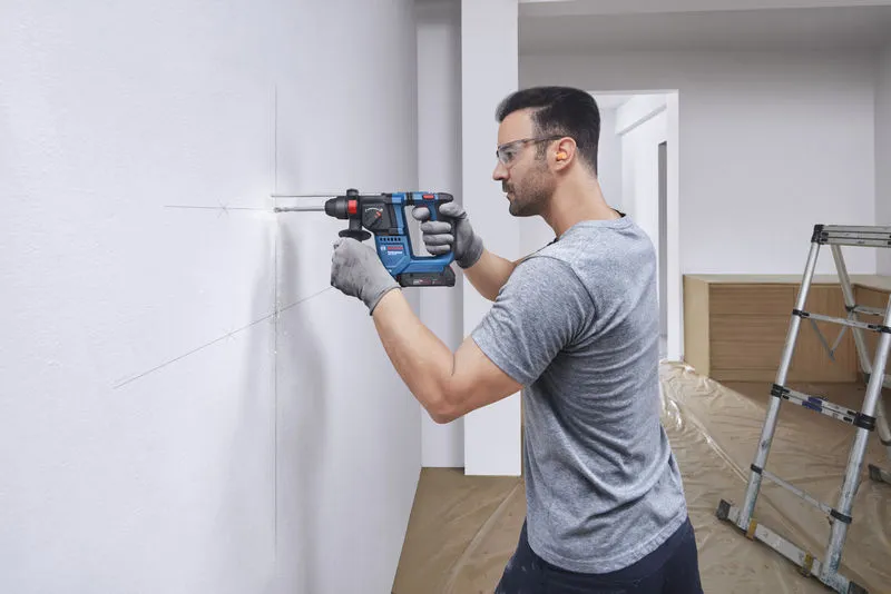 GBH 18V-18 Một người mặc đồ bảo hộ đang khoan lỗ vào bức tường màu trắng được đánh dấu.