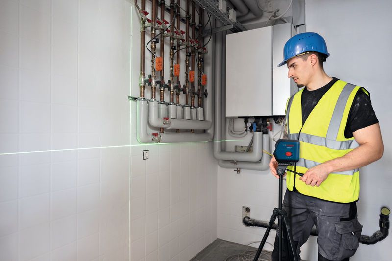 Một người mặc đồ bảo hộ đang lắp đặt máy cân bằng laser gần đường ống trên tường.