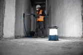 Một công nhân mặc đồ bảo hộ đang khoan tường, đèn công trường không dây trên sàn.