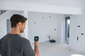 Một người đang quét bức tường trắng bằng camera nhiệt trong một căn phòng chưa hoàn thiện.