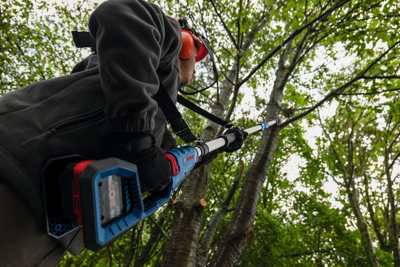 Một người mặc đồ bảo hộ đang cắt tỉa cành cây bằng một dụng cụ điện có thể kéo dài.