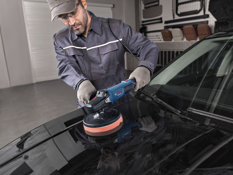 Một người mặc đồ bảo hộ đang đánh bóng mui xe ô tô bằng máy đánh bóng điện.