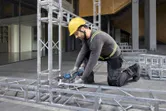 Một người mặc đồ bảo hộ đang siết chặt bu lông trên khung kim loại bằng cờ lê không dây.
