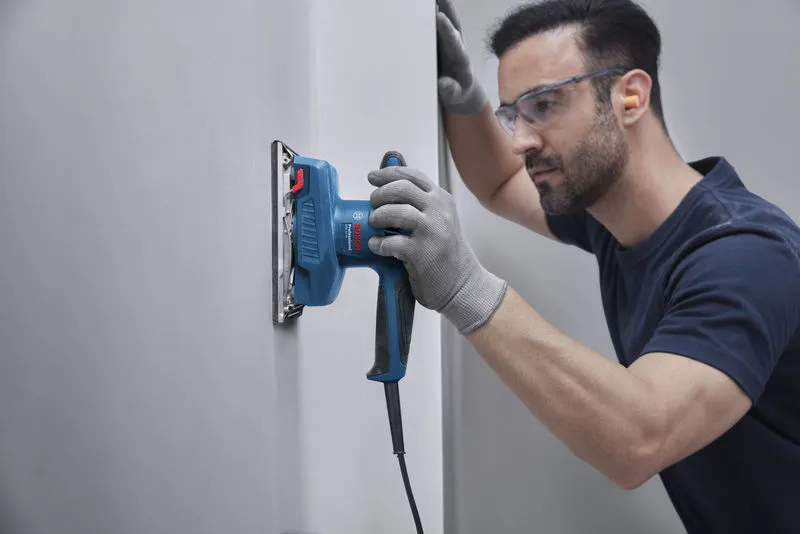 GSS 20-18 Một người mặc đồ bảo hộ đang chà nhám tường bằng máy chà nhám quỹ đạo.