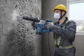 Một người mặc đồ bảo hộ đang sử dụng búa phá dỡ để phá vỡ bức tường bê tông.