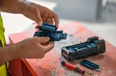Một người đeo thiết bị an toàn đang thay pin cho một dụng cụ điện trên bàn làm việc đầy bụi.