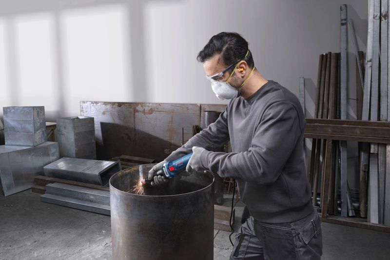 Một người đeo thiết bị an toàn mài cạnh của một thùng kim loại bằng máy mài thẳng.