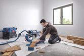 Một người mặc đồ bảo hộ đang cắt ván gỗ bằng máy cưa góc trong một căn phòng sáng.