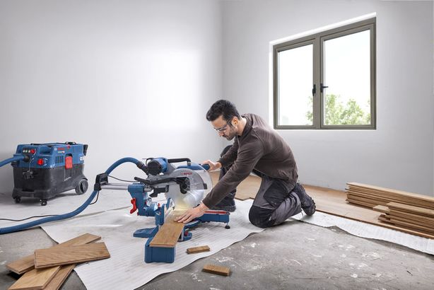 GCM 340-305 D Một người mặc đồ bảo hộ đang cắt ván gỗ bằng máy cưa góc trong một căn phòng sáng.