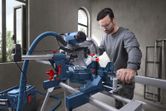 Một người mặc đồ bảo hộ đang cắt gỗ bằng máy cưa góc trong một xưởng.