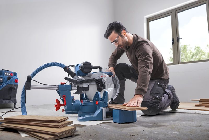 GCM 340-305 D Một người mặc đồ bảo hộ đang cắt sàn gỗ bằng máy cưa góc trong một căn phòng sáng sủa.