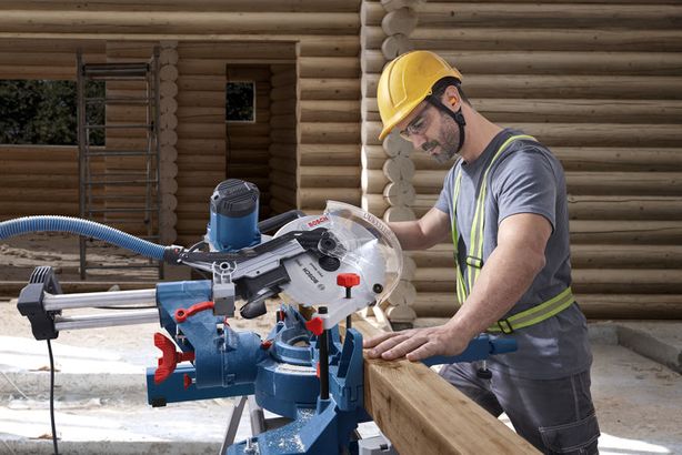 GCM 340-305 D Một người mặc đồ bảo hộ đang cắt gỗ bằng máy cưa góc tại một công trường xây dựng.