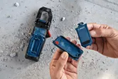 Một người mở ngăn chứa pin màu xanh bên cạnh một dụng cụ trên sàn bê tông bụi bặm.