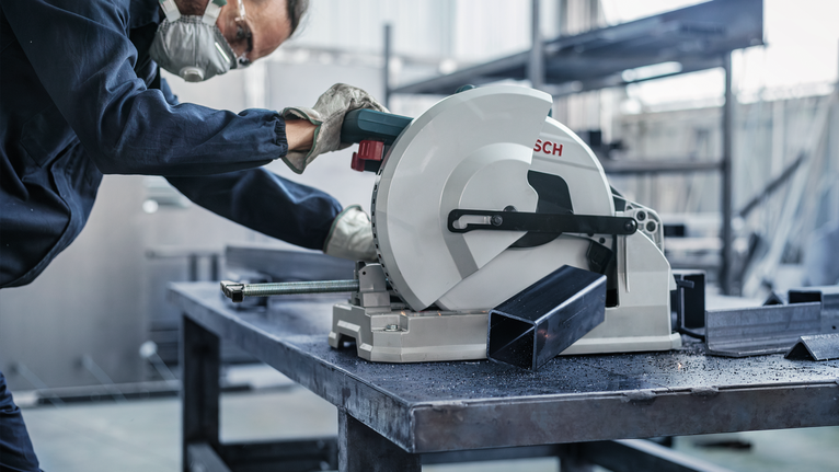 Một công nhân mặc đồ bảo hộ đang cắt một thanh kim loại bằng cưa tròn.