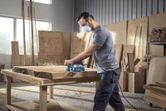 Một người mặc đồ bảo hộ đang bào một tấm gỗ dày trong xưởng mộc.