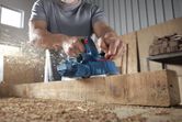 Một người mặc đồ bảo hộ đang di chuyển một thanh gỗ, làm tung bay nhiều mảnh gỗ vụn.