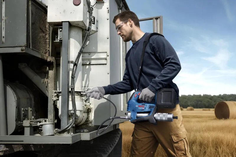 Một người mặc đồ bảo hộ sử dụng súng bơm mỡ không dây để bảo dưỡng máy móc nông nghiệp.