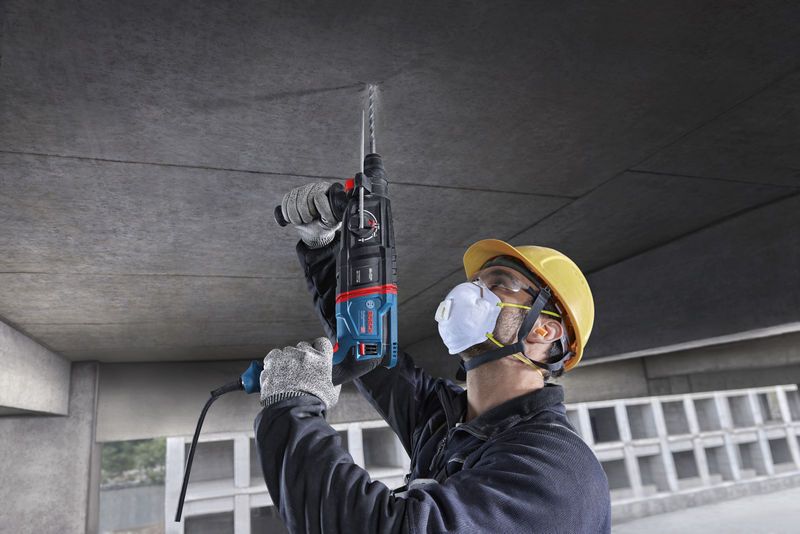 Một người mặc đồ bảo hộ đang khoan vào trần bê tông bằng búa quay.