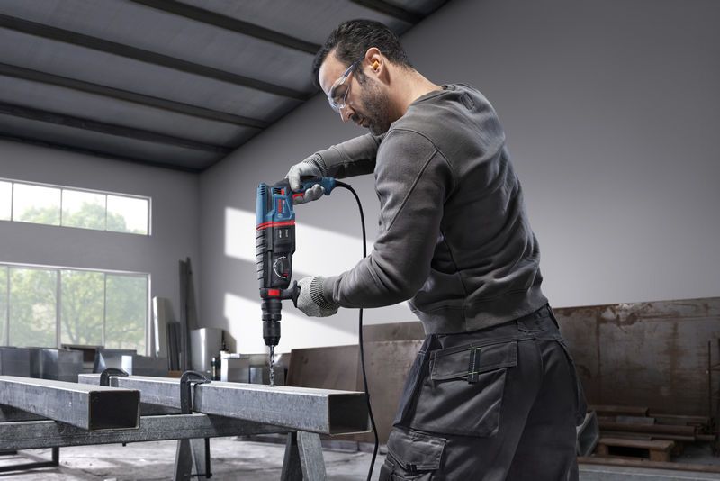 Một người mặc đồ bảo hộ đang khoan vào một thanh thép bằng búa quay.