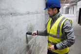 Một người mặc thiết bị an toàn đang kiểm tra độ ẩm của tường bê tông bằng máy đo độ ẩm vật liệu.