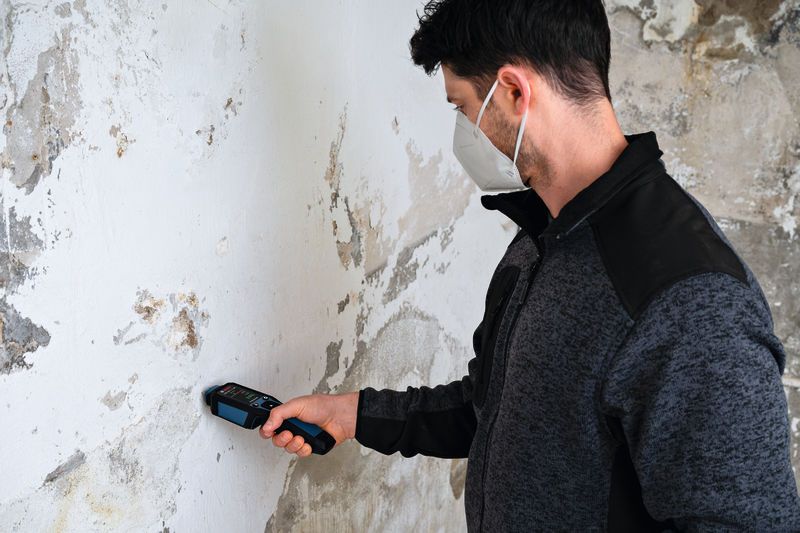 Một người mặc đồ bảo hộ đang sử dụng máy đo độ ẩm vật liệu trên bức tường bị hư hỏng.