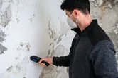 Một người mặc đồ bảo hộ đang sử dụng máy đo độ ẩm vật liệu trên bức tường bị hư hỏng.